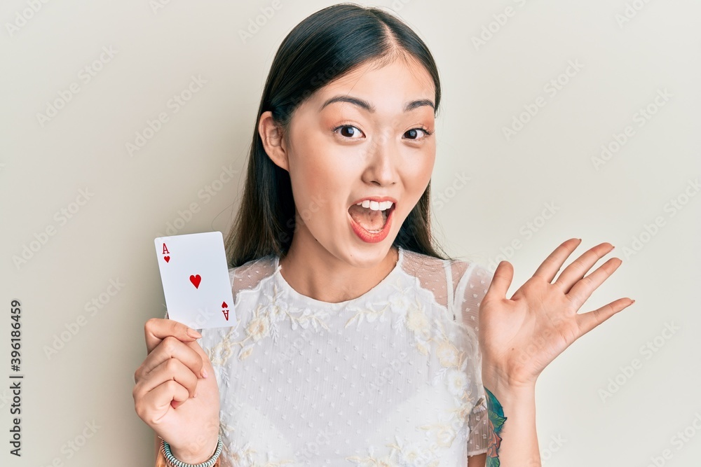 Young chinese woman holding ace poker card celebrating achievement with happy smile and winner expression with raised hand