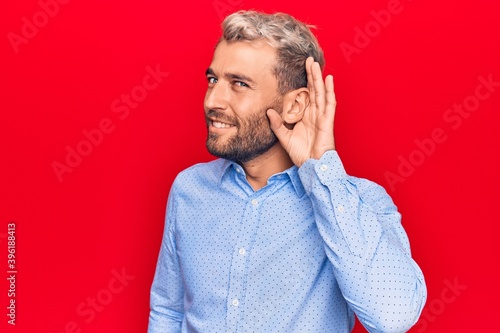 Young handsome blond man wearing casual shirt standing over isolated red background smiling with hand over ear listening and hearing to rumor or gossip. Deafness concept.