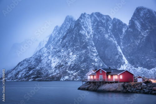 Fotografie Red rorbu on sea coast and snow covered mountain in fog at dusk