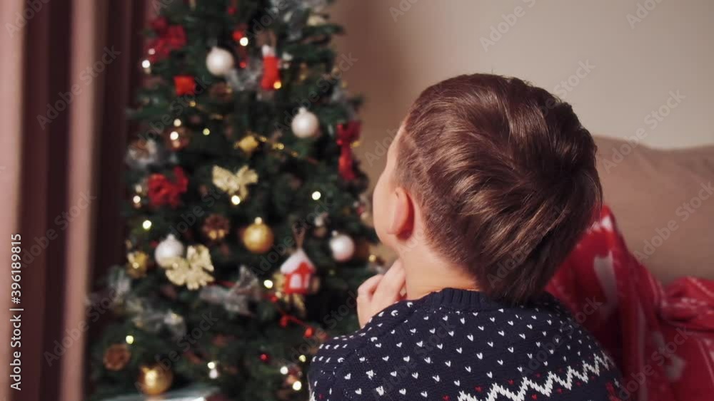 A dark-haired boy at home looks at the Christmas tree, hands folded together. The boy prays and hopes for the best. Celebrating Christmas at home. A dark-haired boy at home looks at the Christmas tree, hands folded together. The boy prays and hopes for the best. Celebrating Christmas at home.