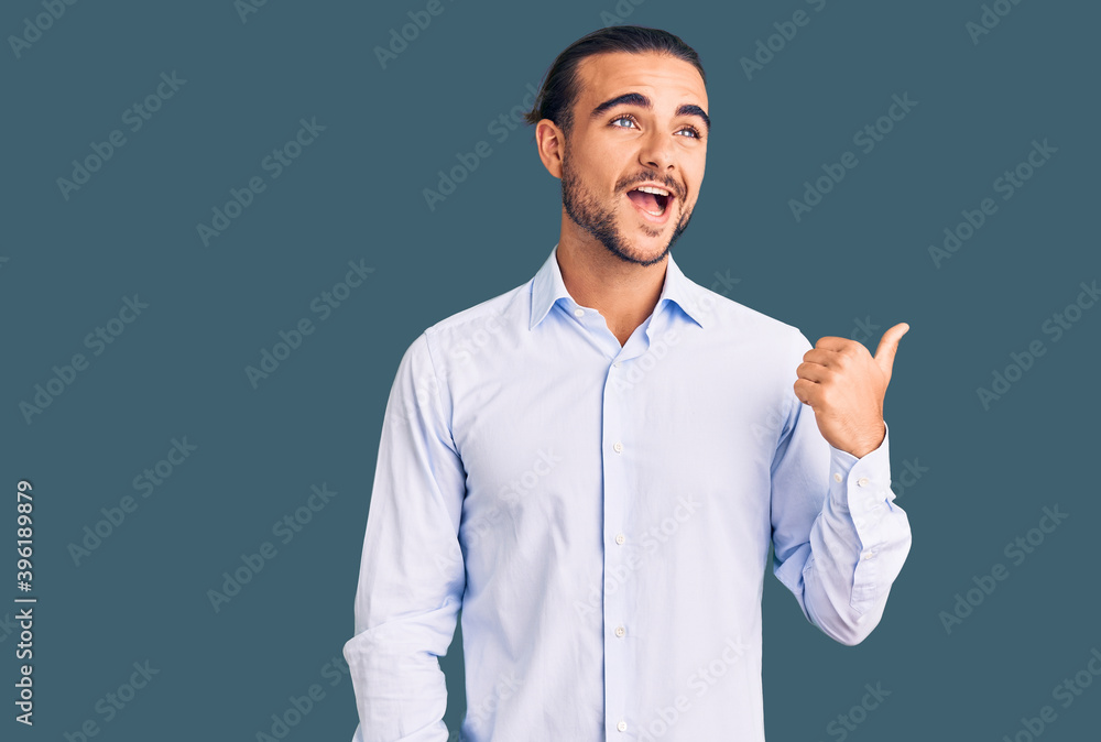 Young handsome man wearing business clothes smiling with happy face looking and pointing to the side with thumb up. Young handsome man wearing business clothes smiling with happy face looking and pointing to the side with thumb up.