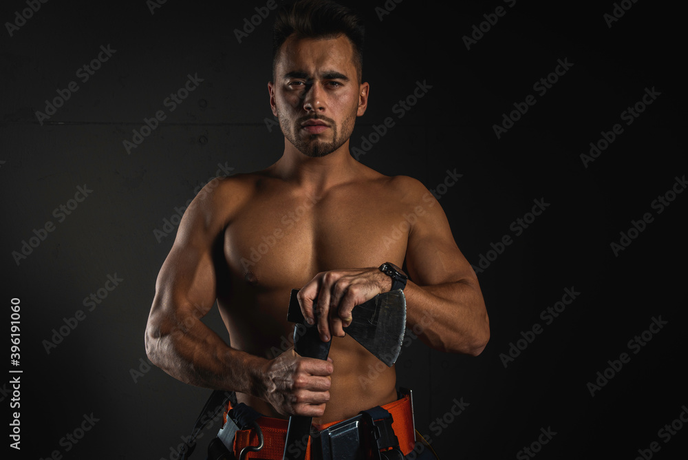 young handsome adult, muscular firefighter in uniform holding an ax of fire equipment in his hands in front of him, pensively, isolated on a dark background. Low key. Protection concept