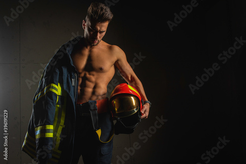 Murais de parede young handsome adult, muscular firefighter in uniform holding ax of fire equipment in his hands, pensive, isolated on dark background