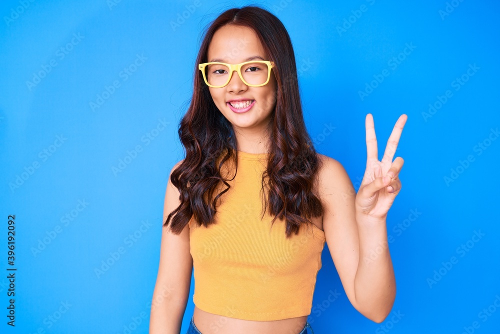 Young beautiful chinese girl wearing casual clothes and glasses smiling with happy face winking at the camera doing victory sign. number two. Young beautiful chinese girl wearing casual clothes and glasses smiling with happy face winking at the camera doing victory sign. number two.