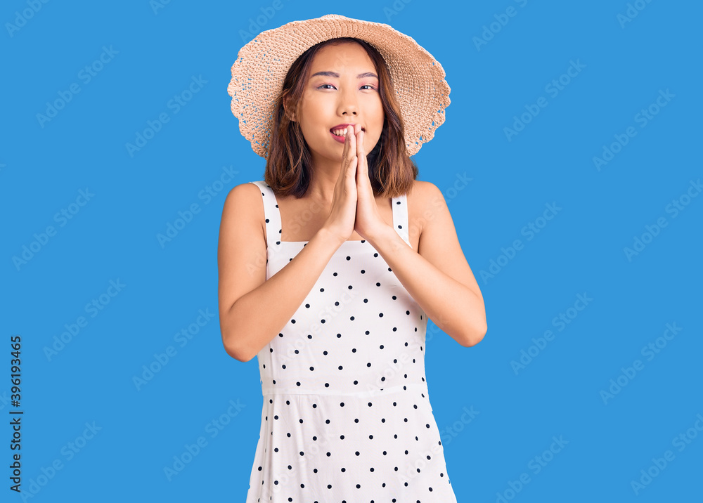 Young beautiful chinese girl wearing summer hat praying with hands together asking for forgiveness smiling confident.