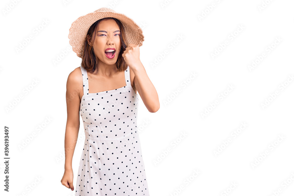 Young beautiful chinese girl wearing summer hat angry and mad raising fist frustrated and furious while shouting with anger. rage and aggressive concept. Young beautiful chinese girl wearing summer hat angry and mad raising fist frustrated and furious while shouting with anger. rage and aggressive concept.