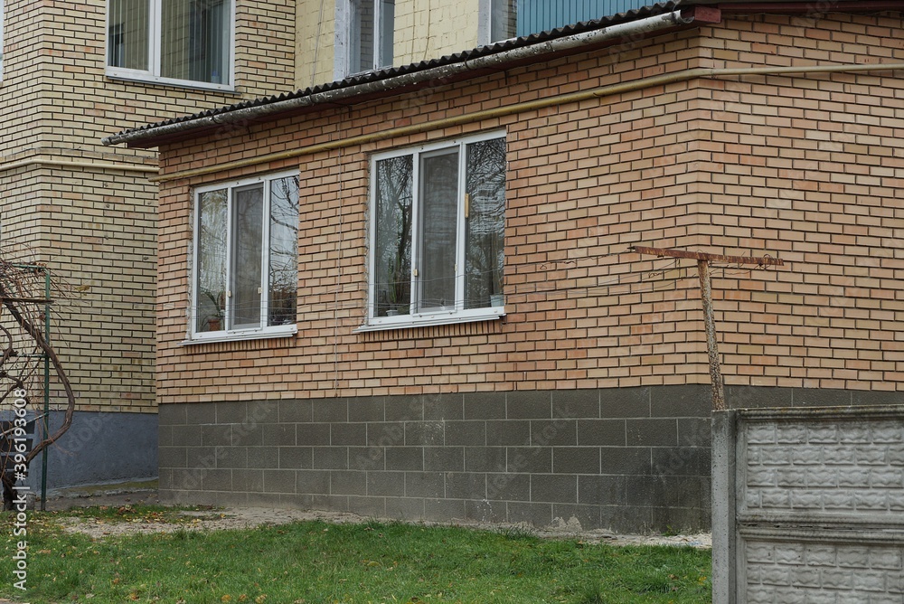 facade of a brown brick house with two windows and a black foundation on the street in green grass