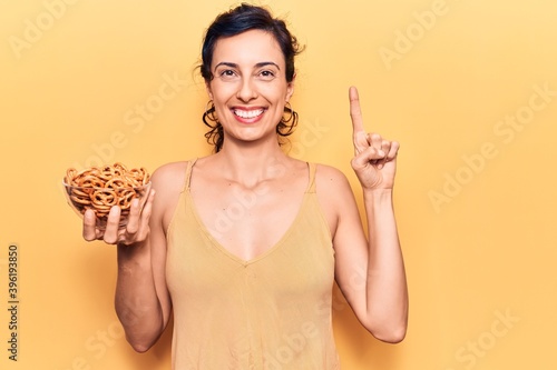 Young beautiful hispanic woman holding pretzels smiling with an idea or question pointing finger with happy face  number one