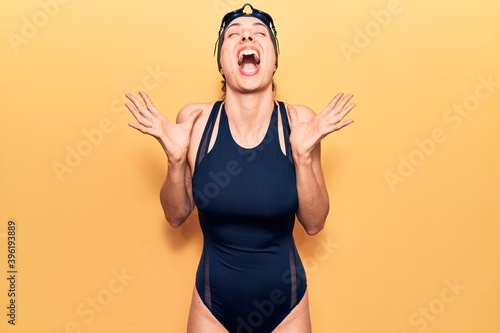 Young beautiful hispanic woman wearing swimwear and swimmer glasses celebrating mad and crazy for success with arms raised and closed eyes screaming excited. winner concept