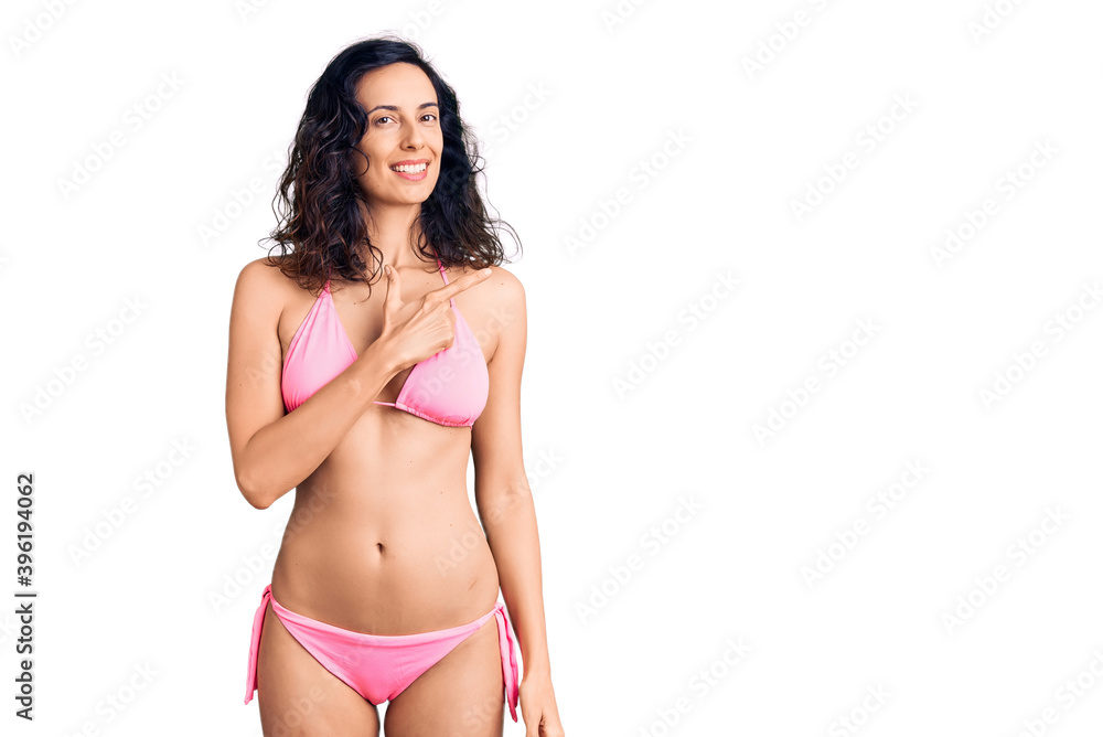 Naklejka premium Young beautiful hispanic woman wearing bikini cheerful with a smile on face pointing with hand and finger up to the side with happy and natural expression