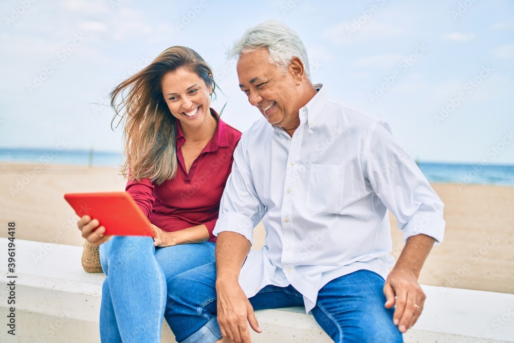 Middle age hispanic couple using touchpad sitting on the bench at the beach.