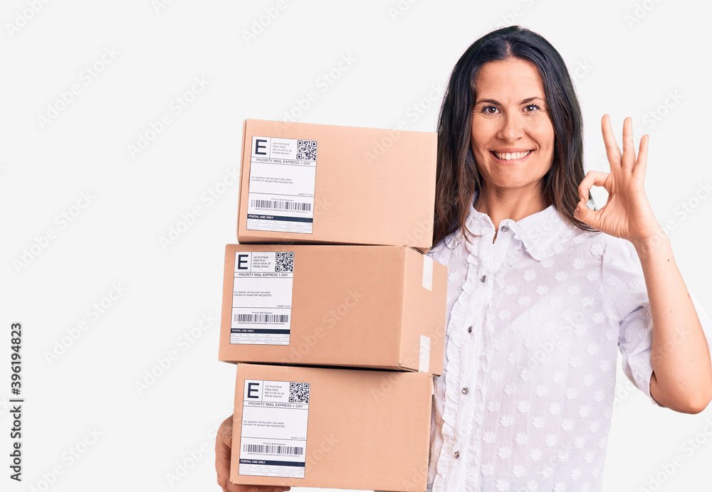 Young beautiful brunette woman holding delivery cardboard package doing ok sign with fingers, smiling friendly gesturing excellent symbol Young beautiful brunette woman holding delivery cardboard package doing ok sign with fingers, smiling friendly gesturing excellent symbol