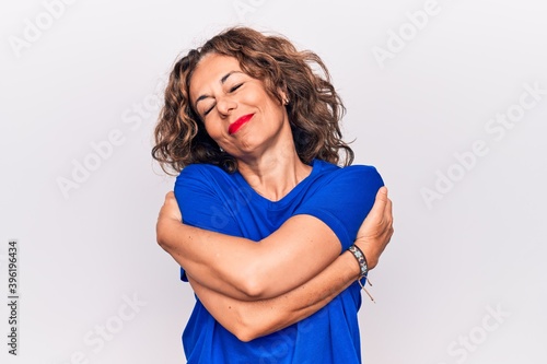 Middle age beautiful brunette woman wearing blue t-shirt standing over white background hugging oneself happy and positive, smiling confident. Self love and self care