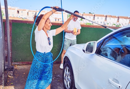 Middle age beautiful couple wearing casual clothes and smiling happy. Standing with smile on face washing car using water pistol and sponge.