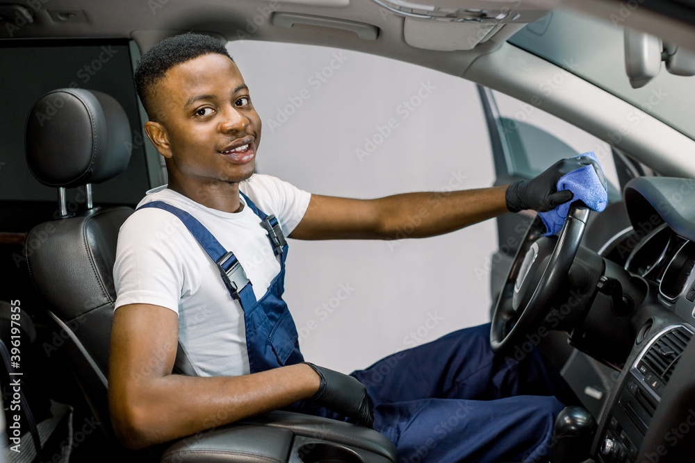 Handsome African young man in uniform and protective gloves, cleaning car interior and steering wheel using microfiber cloth, smiling at camera. Car detailing or valeting concept. Selective focus Handsome African young man in uniform and protective gloves, cleaning car interior and steering wheel using microfiber cloth, smiling at camera. Car detailing or valeting concept. Selective focus