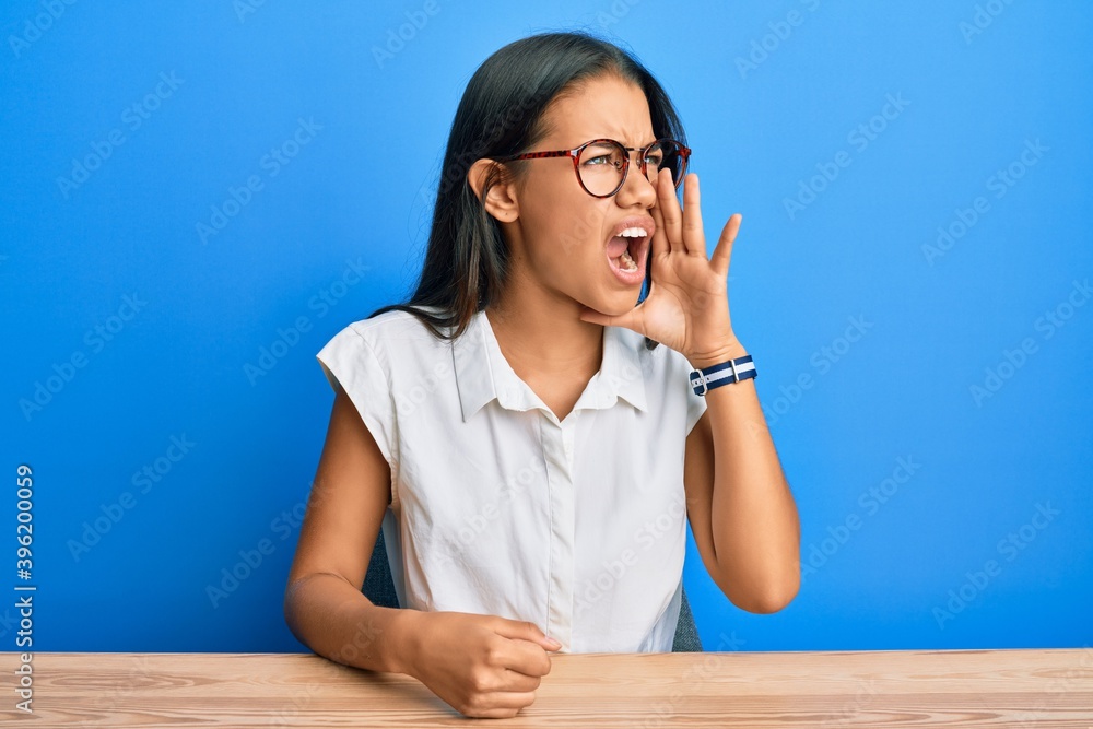 Beautiful hispanic woman wearing casual clothes sitting on the table shouting and screaming loud to side with hand on mouth. communication concept. Beautiful hispanic woman wearing casual clothes sitting on the table shouting and screaming loud to side with hand on mouth. communication concept.