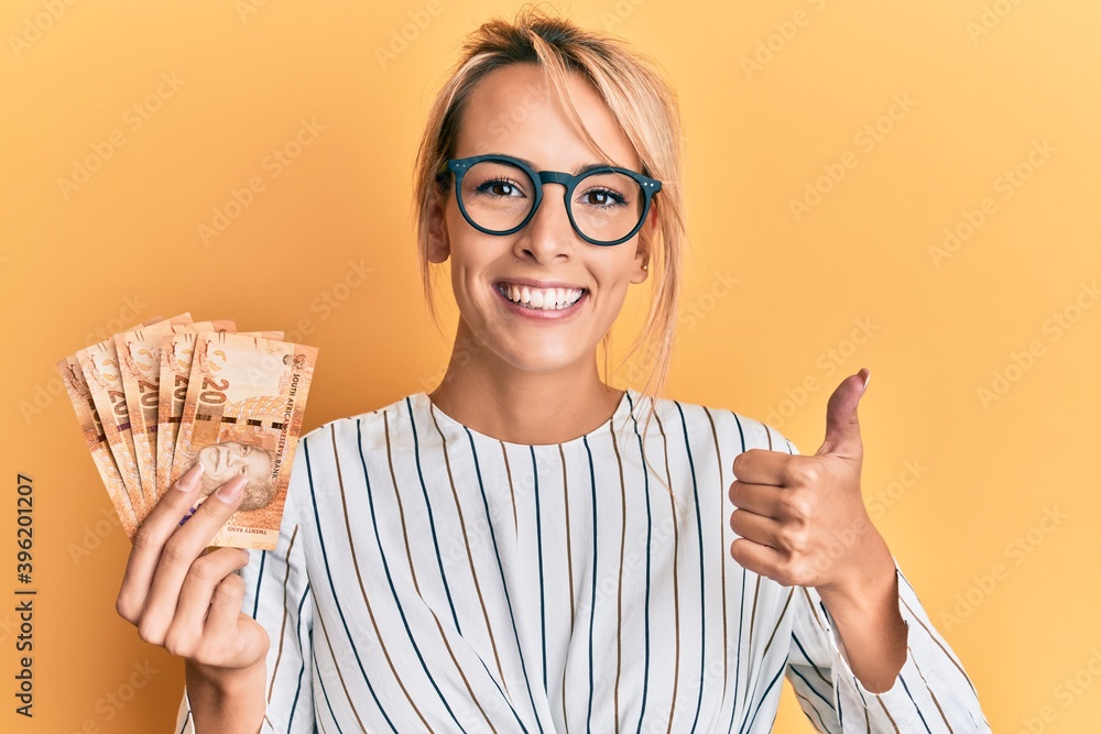 Beautiful blonde woman holding south african 20 rand banknotes smiling happy and positive, thumb up doing excellent and approval sign