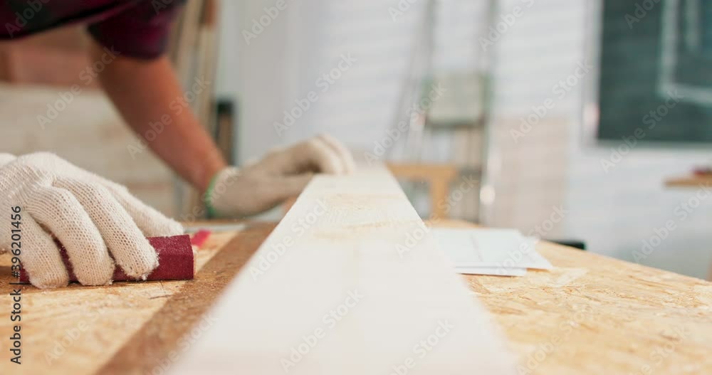 A bearded handyman is sanding wood for furniture in a carpentry workshop. The carpenter blows off the chips after sanding the board. A bearded handyman is sanding wood for furniture in a carpentry workshop. The carpenter blows off the chips after sanding the board.