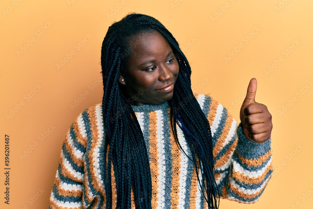 Young black woman with braids wearing casual winter sweater looking proud, smiling doing thumbs up gesture to the side Young black woman with braids wearing casual winter sweater looking proud, smiling doing thumbs up gesture to the side