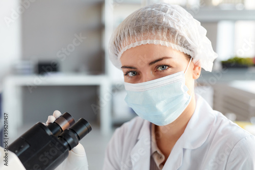 Close up portrait of young female scientist wearing face mask and looking at camera while working with microscope in medical laboratory, copy space above