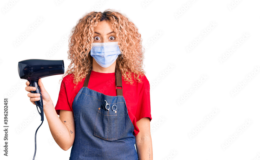 Young blonde woman with curly hair holding dryer blow wearing safety mask for coranvirus scared and amazed with open mouth for surprise, disbelief face Young blonde woman with curly hair holding dryer blow wearing safety mask for coranvirus scared and amazed with open mouth for surprise, disbelief face