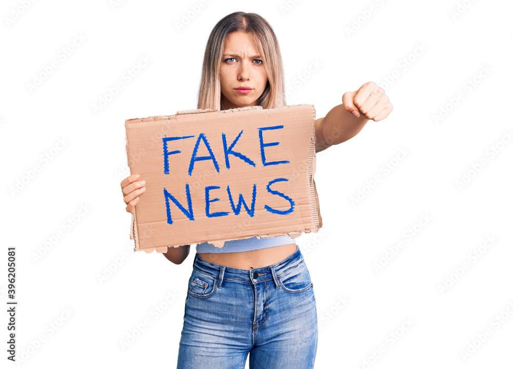 Young beautiful blonde woman holding fake news banner annoyed and frustrated shouting with anger, yelling crazy with anger and hand raised Young beautiful blonde woman holding fake news banner annoyed and frustrated shouting with anger, yelling crazy with anger and hand raised