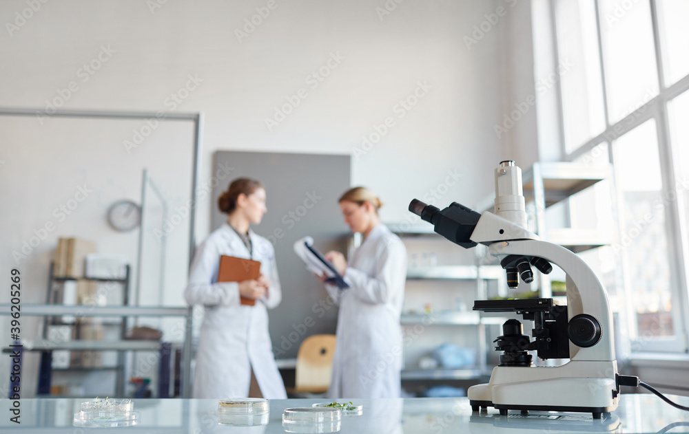 Naklejka premium Portrait of two female scientists discussing research while standing in medical laboratory, focus on microscope in foreground, copy space