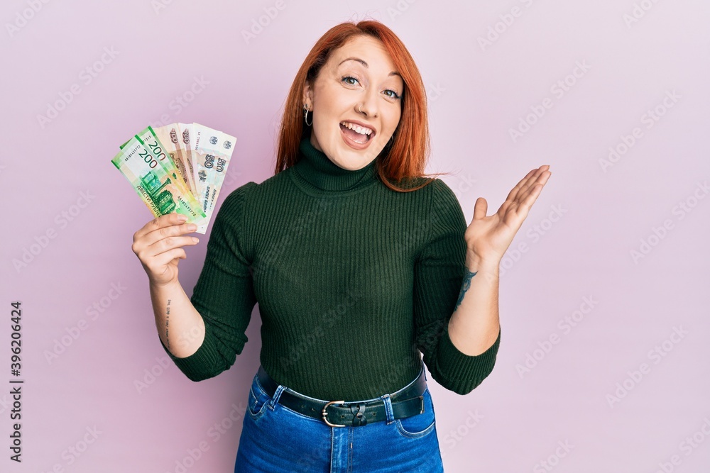 Beautiful redhead woman holding russian ruble banknotes celebrating achievement with happy smile and winner expression with raised hand Beautiful redhead woman holding russian ruble banknotes celebrating achievement with happy smile and winner expression with raised hand