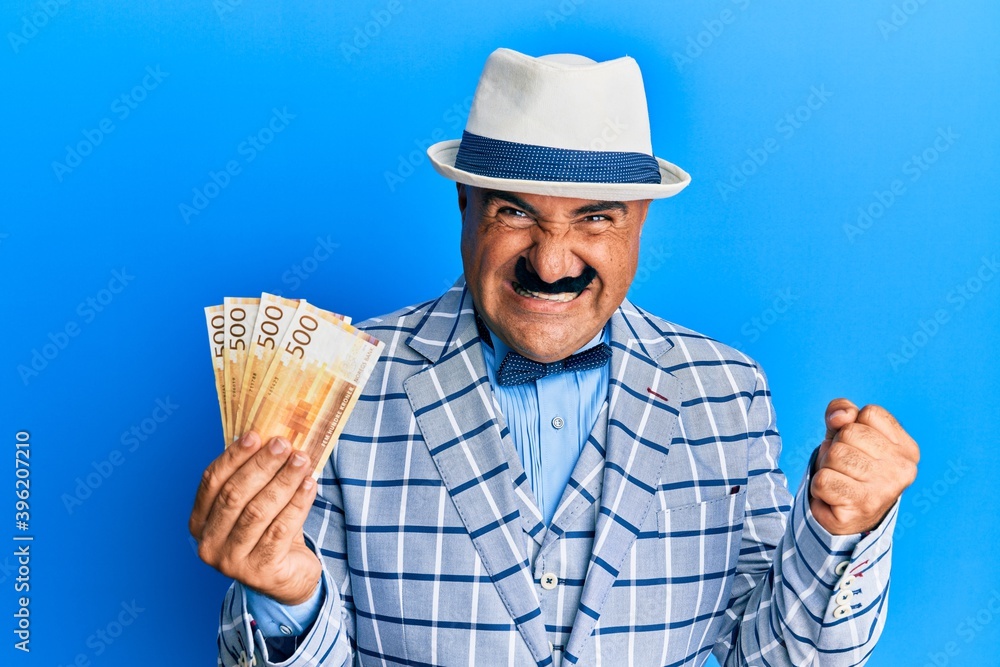 Mature middle east man with mustache wearing elegant vintage style holding norwegian krone screaming proud, celebrating victory and success very excited with raised arm Mature middle east man with mustache wearing elegant vintage style holding norwegian krone screaming proud, celebrating victory and success very excited with raised arm