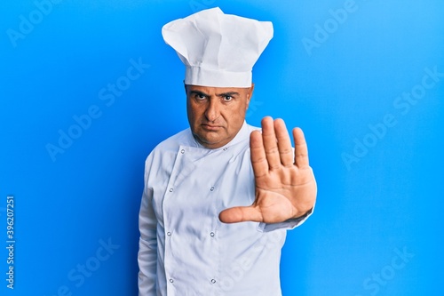 Mature middle east man wearing professional cook uniform and hat doing stop sing with palm of the hand. warning expression with negative and serious gesture on the face.
