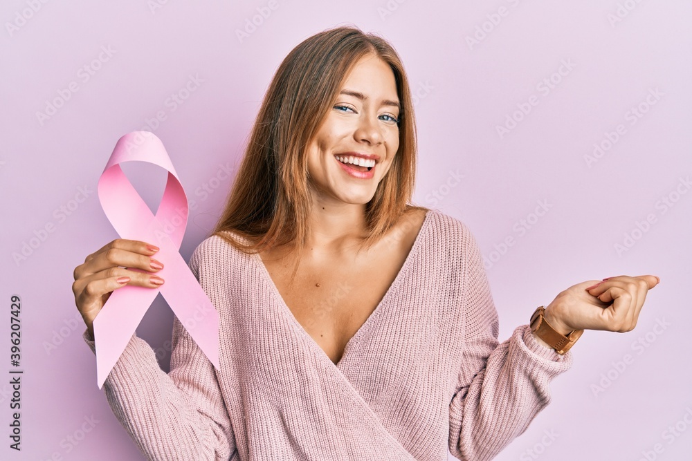Beautiful young blonde woman holding pink cancer ribbon screaming proud, celebrating victory and success very excited with raised arm Beautiful young blonde woman holding pink cancer ribbon screaming proud, celebrating victory and success very excited with raised arm
