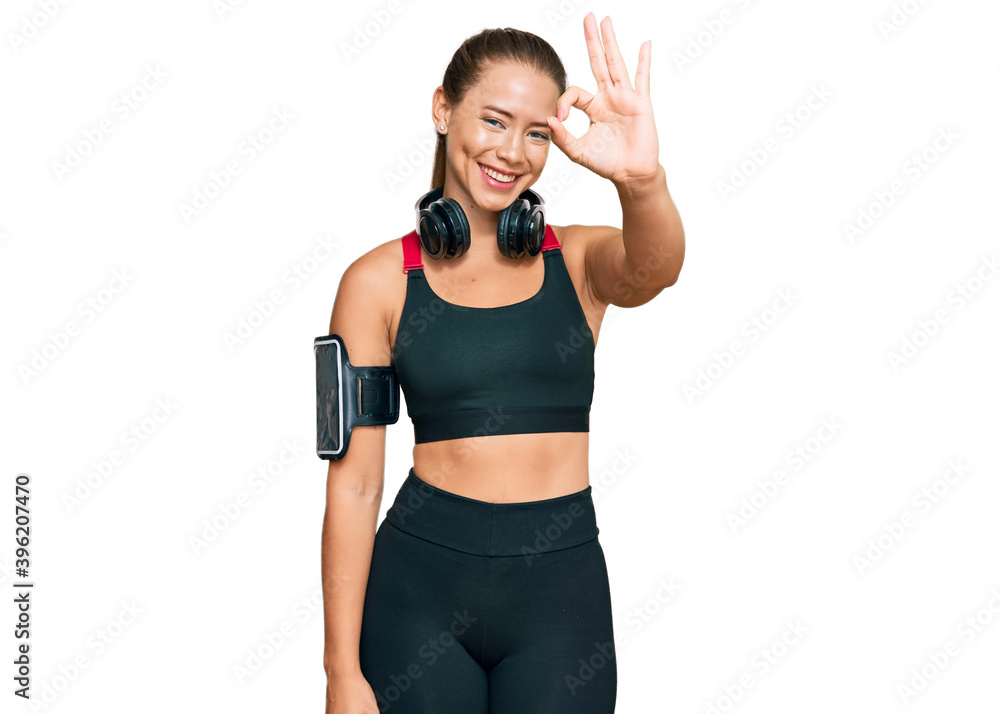 Beautiful blonde woman wearing gym clothes and using headphones smiling positive doing ok sign with hand and fingers. successful expression. Beautiful blonde woman wearing gym clothes and using headphones smiling positive doing ok sign with hand and fingers. successful expression.