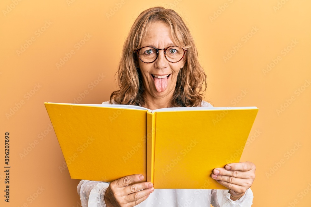 Middle age blonde woman reading a book wearing glasses sticking tongue out happy with funny expression. Middle age blonde woman reading a book wearing glasses sticking tongue out happy with funny expression.