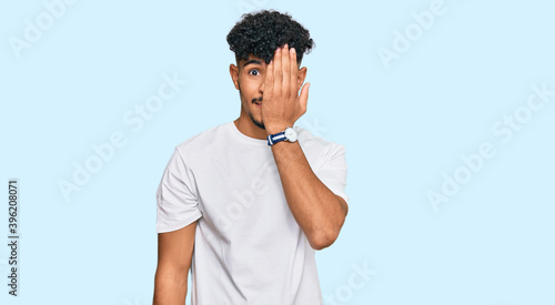 Young arab man wearing casual white t shirt covering one eye with hand, confident smile on face and surprise emotion.