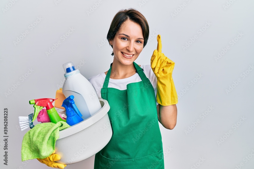 Young brunette woman with short hair wearing apron holding cleaning products pointing finger up with successful idea. exited and happy. number one. Young brunette woman with short hair wearing apron holding cleaning products pointing finger up with successful idea. exited and happy. number one.