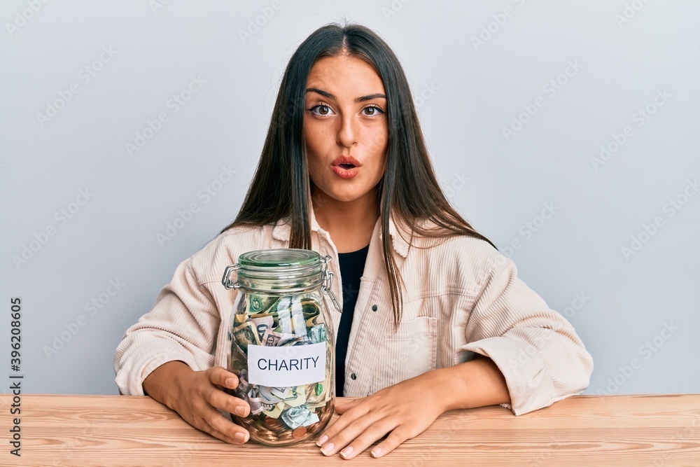 Beautiful hispanic woman holding charity jar with money scared and amazed with open mouth for surprise, disbelief face Beautiful hispanic woman holding charity jar with money scared and amazed with open mouth for surprise, disbelief face