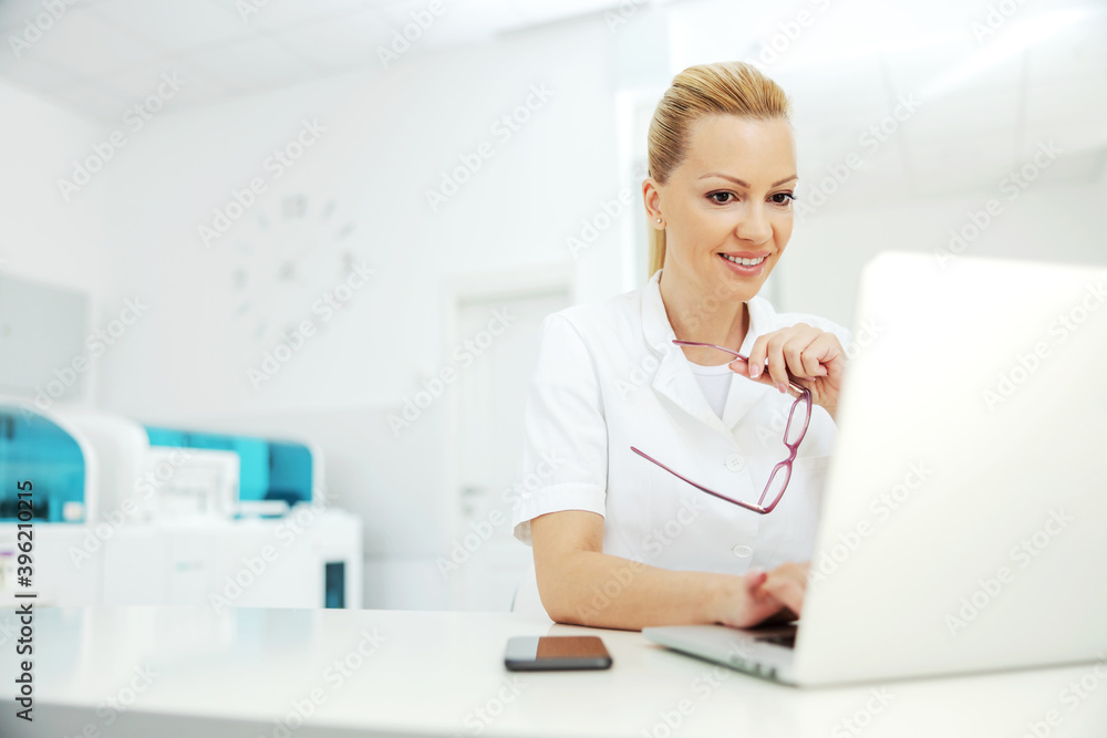 Portrait of handsome smiling blond female laboratory assistant sitting in lab and using laptop for entering test results. Portrait of handsome smiling blond female laboratory assistant sitting in lab and using laptop for entering test results.