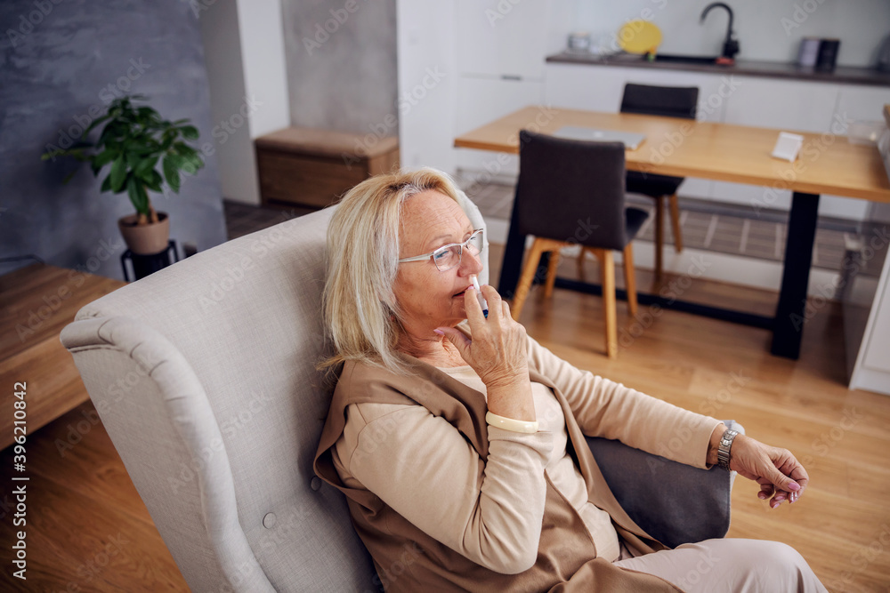 Blond senior woman sitting in chair at home and spraying nose spray. Blond senior woman sitting in chair at home and spraying nose spray.
