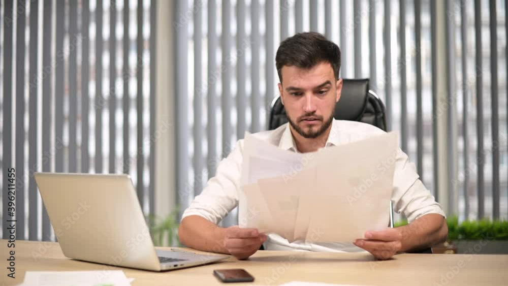 The young head of the company examines the reports printed on paper and is perplexed. Young boss is not happy with this season's performance The young head of the company examines the reports printed on paper and is perplexed. Young boss is not happy with this season's performance