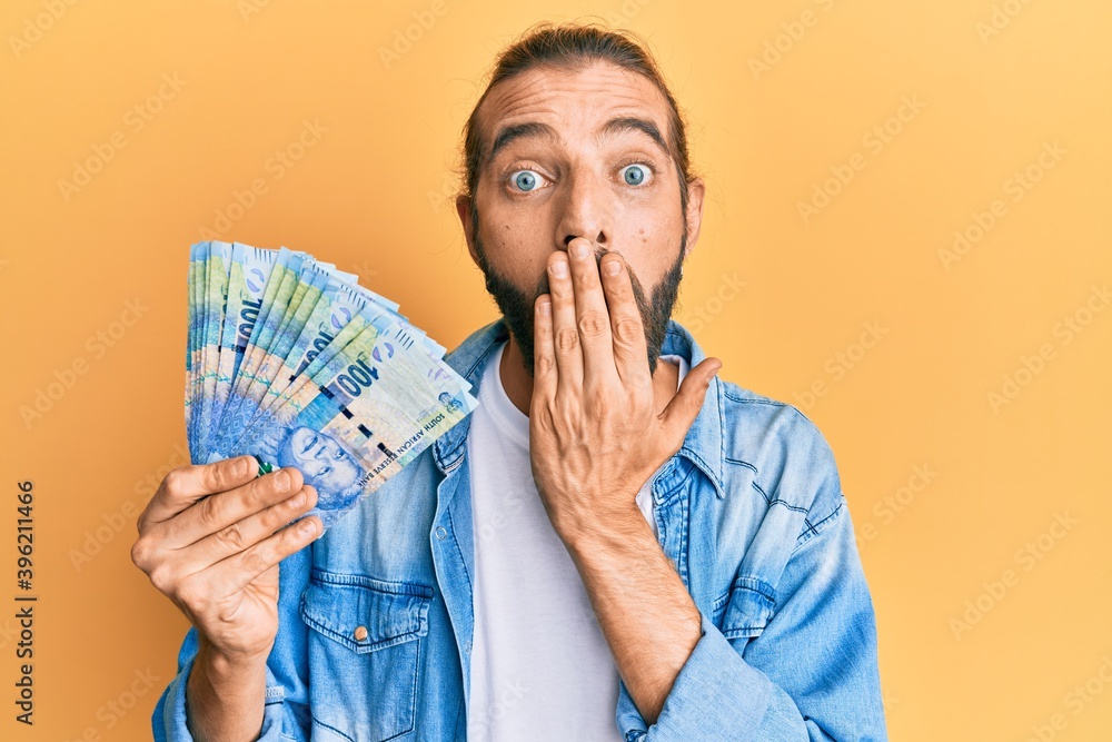 Attractive man with long hair and beard holding south african 100 rand banknotes covering mouth with hand, shocked and afraid for mistake. surprised expression Attractive man with long hair and beard holding south african 100 rand banknotes covering mouth with hand, shocked and afraid for mistake. surprised expression