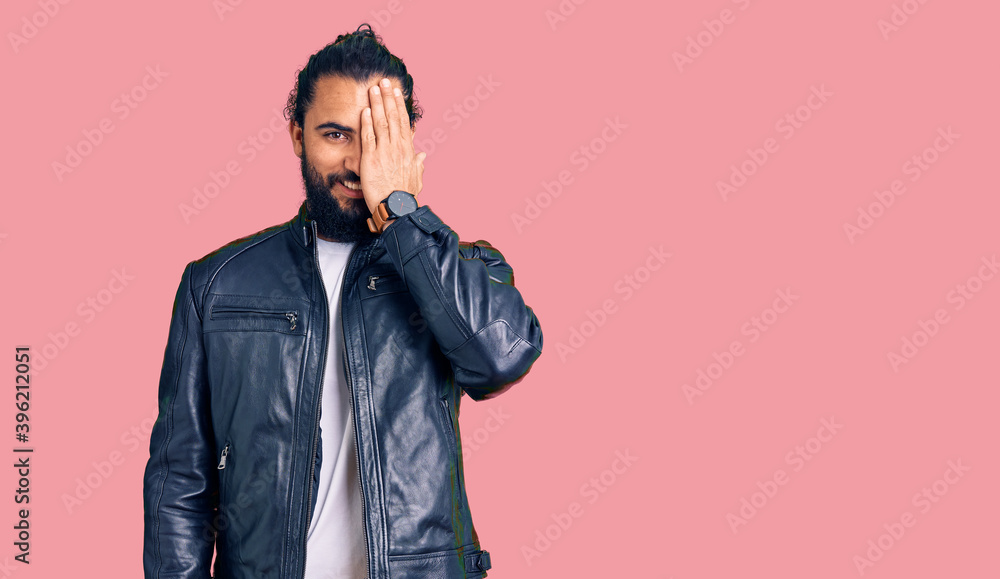 Young arab man wearing casual leather jacket covering one eye with hand, confident smile on face and surprise emotion. Young arab man wearing casual leather jacket covering one eye with hand, confident smile on face and surprise emotion.