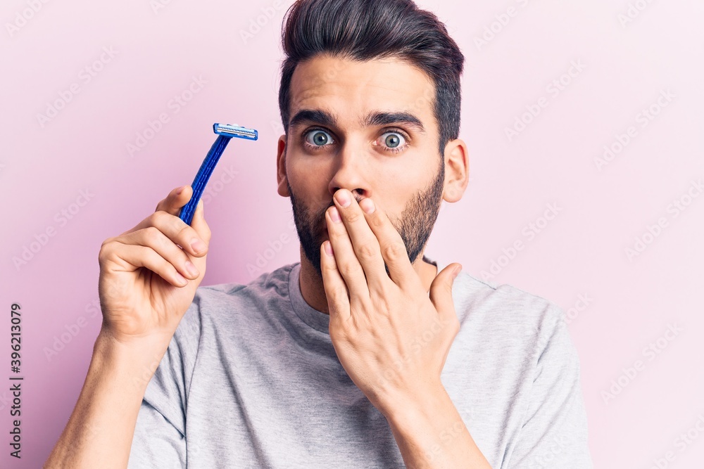 Young handsome man with beard holding razor covering mouth with hand, shocked and afraid for mistake. surprised expression Young handsome man with beard holding razor covering mouth with hand, shocked and afraid for mistake. surprised expression
