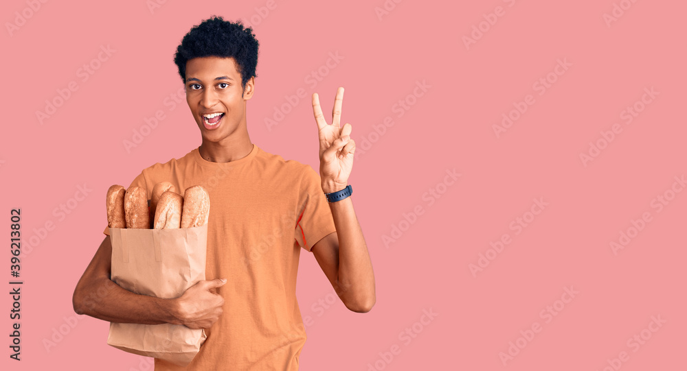 Young african american man holding paper bag with bread smiling looking to the camera showing fingers doing victory sign. number two. Young african american man holding paper bag with bread smiling looking to the camera showing fingers doing victory sign. number two.