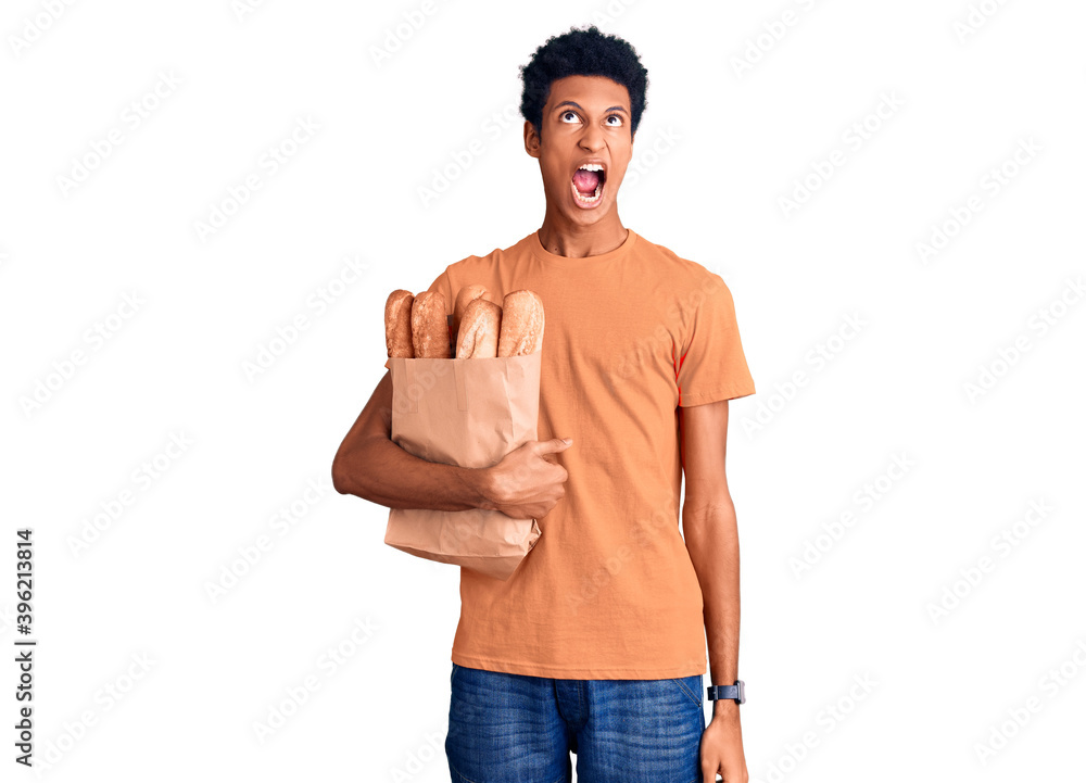 Young african american man holding paper bag with bread angry and mad screaming frustrated and furious, shouting with anger. rage and aggressive concept. Young african american man holding paper bag with bread angry and mad screaming frustrated and furious, shouting with anger. rage and aggressive concept.