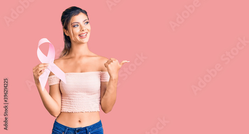Young beautiful woman holding cancer awareness pink ribbon pointing thumb up to the side smiling happy with open mouth