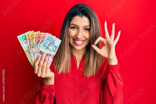 Beautiful brunette woman holding australian dollars smiling positive doing ok sign with hand and fingers. successful expression. photo