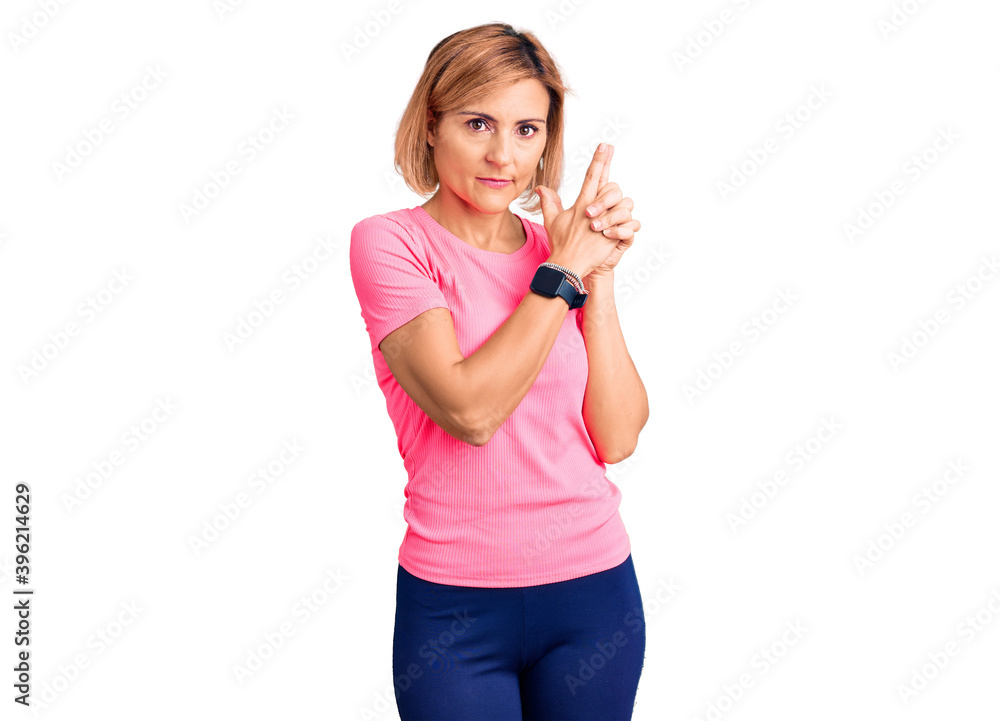 Young blonde woman wearing sportswear holding symbolic gun with hand gesture, playing killing shooting weapons, angry face Young blonde woman wearing sportswear holding symbolic gun with hand gesture, playing killing shooting weapons, angry face