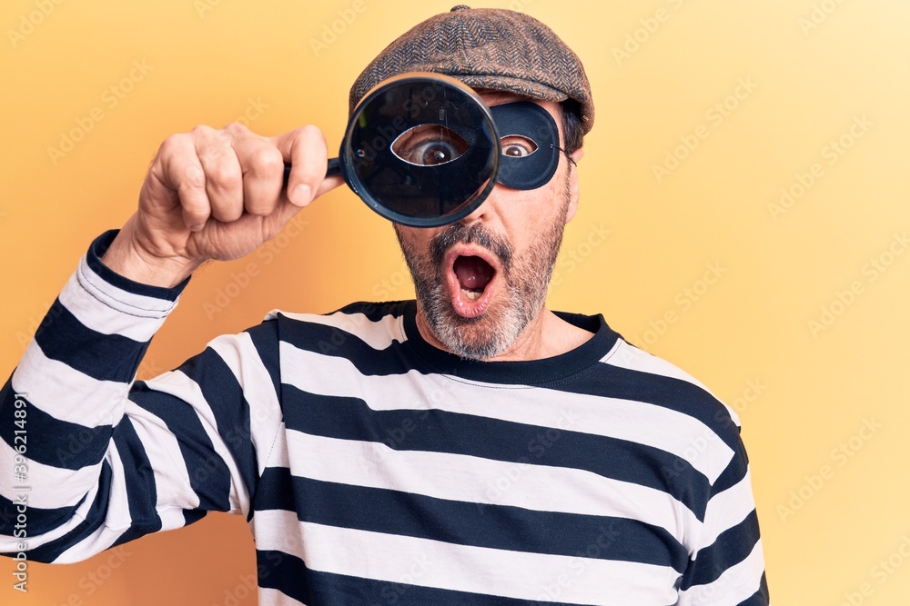 Middle age handsome burglar man searching using magnifying glass over yellow background scared and amazed with open mouth for surprise, disbelief face Middle age handsome burglar man searching using magnifying glass over yellow background scared and amazed with open mouth for surprise, disbelief face