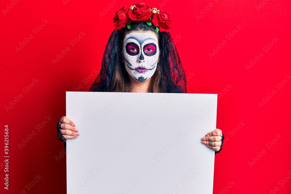 Young woman wearing day of the dead costume holding blank empty banner thinking attitude and sober expression looking self confident Young woman wearing day of the dead costume holding blank empty banner thinking attitude and sober expression looking self confident