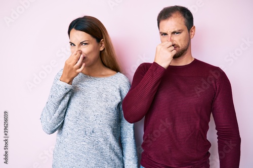 Beautiful couple wearing casual clothes smelling something stinky and disgusting, intolerable smell, holding breath with fingers on nose. bad smell